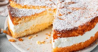 Angeschnittene Käsesahnetorte auf einer Tortenplatte, dahinter Sahne und eine Zitrone