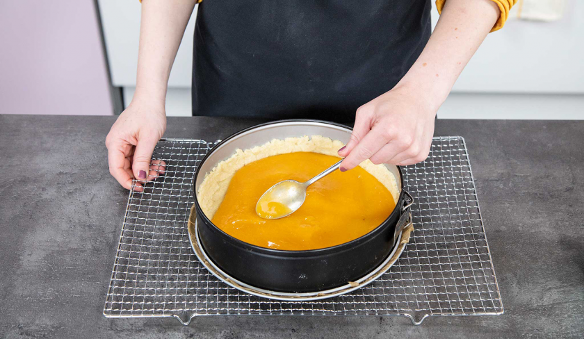 Aprikosenpüree wird auf dem Boden für den Aprikosenkuchen mit Streuseln mit einem Löffel verstrichen.