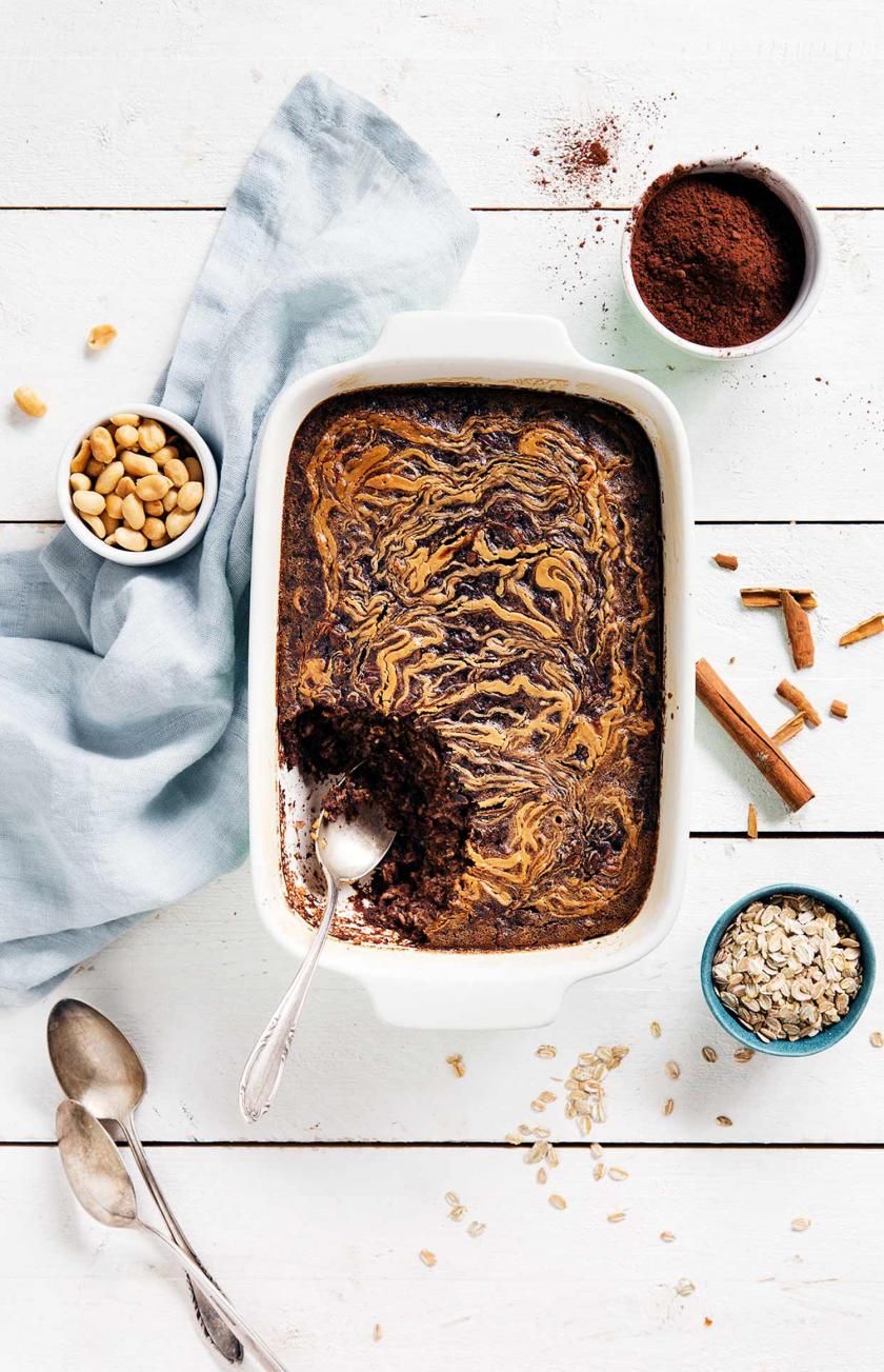 Baked Oatmeal auf einem Tisch. Drumherum Nüsse, Haferflocken.