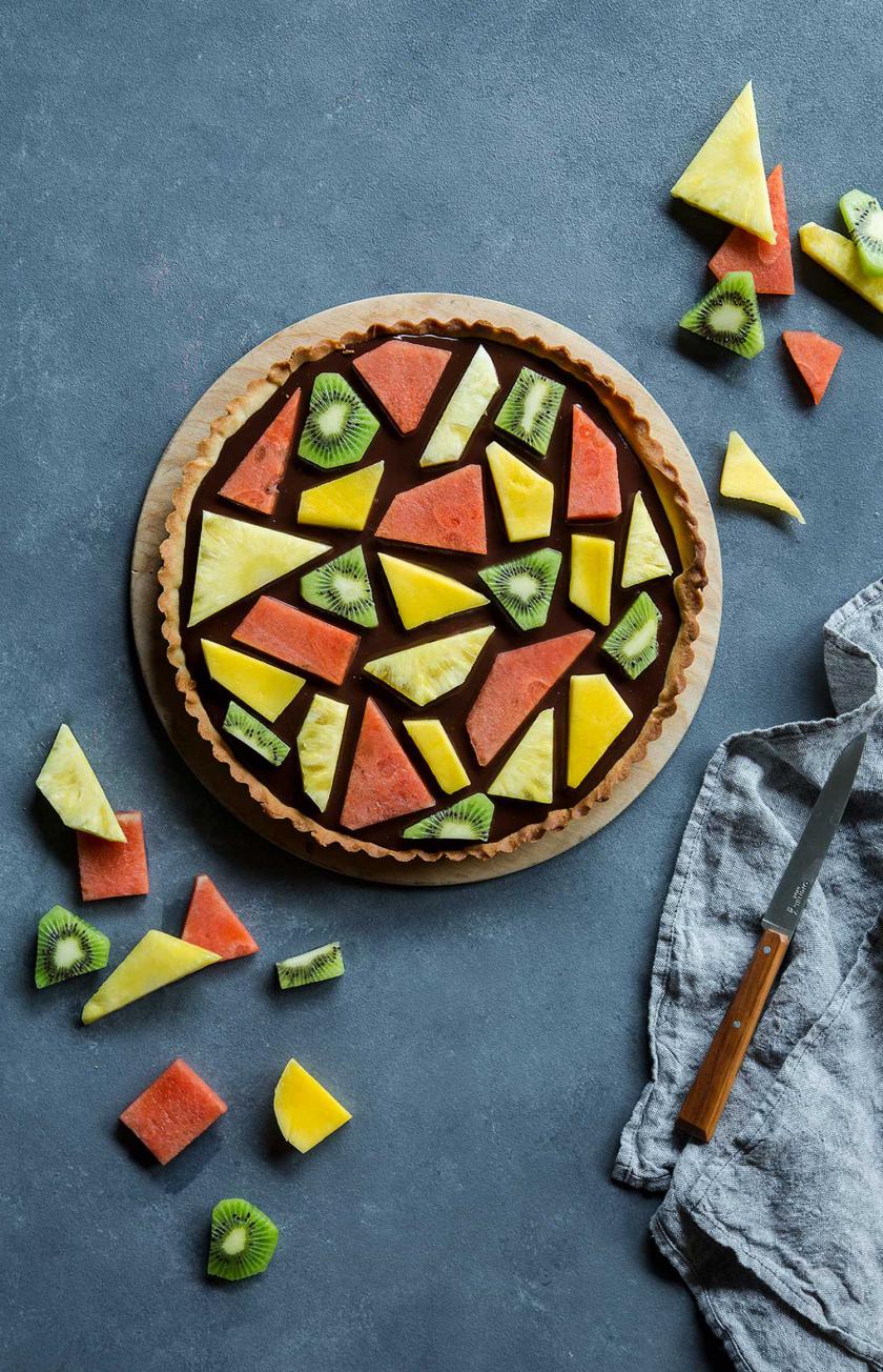 Fruchtiger Mosaik Kuchen auf einem Holzteller angerichtet. Obst drumherum.