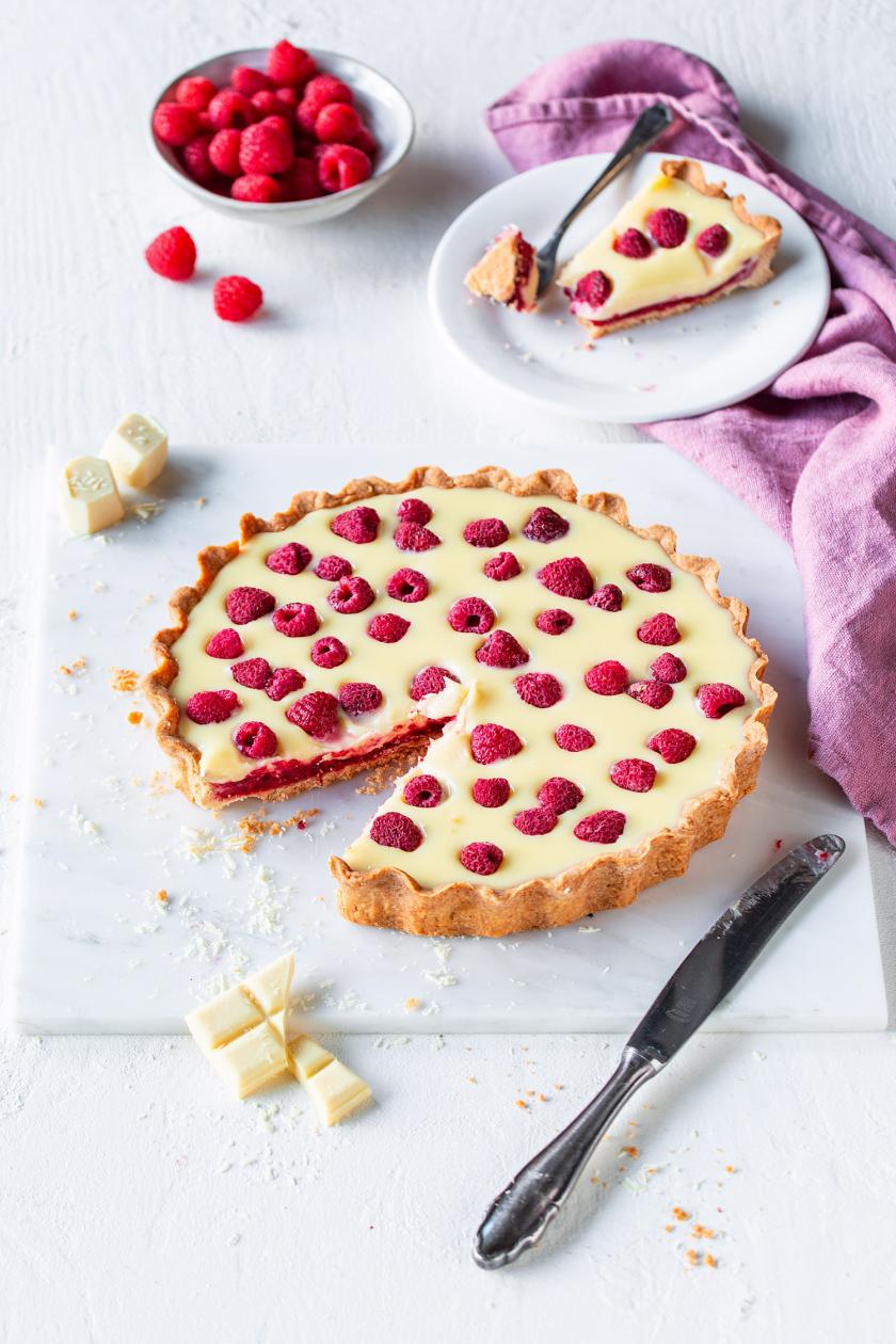 Himbeertarte mit weißer Schokolade angeschnitten auf einer Kuchenplatte mit angelegtem Messer.