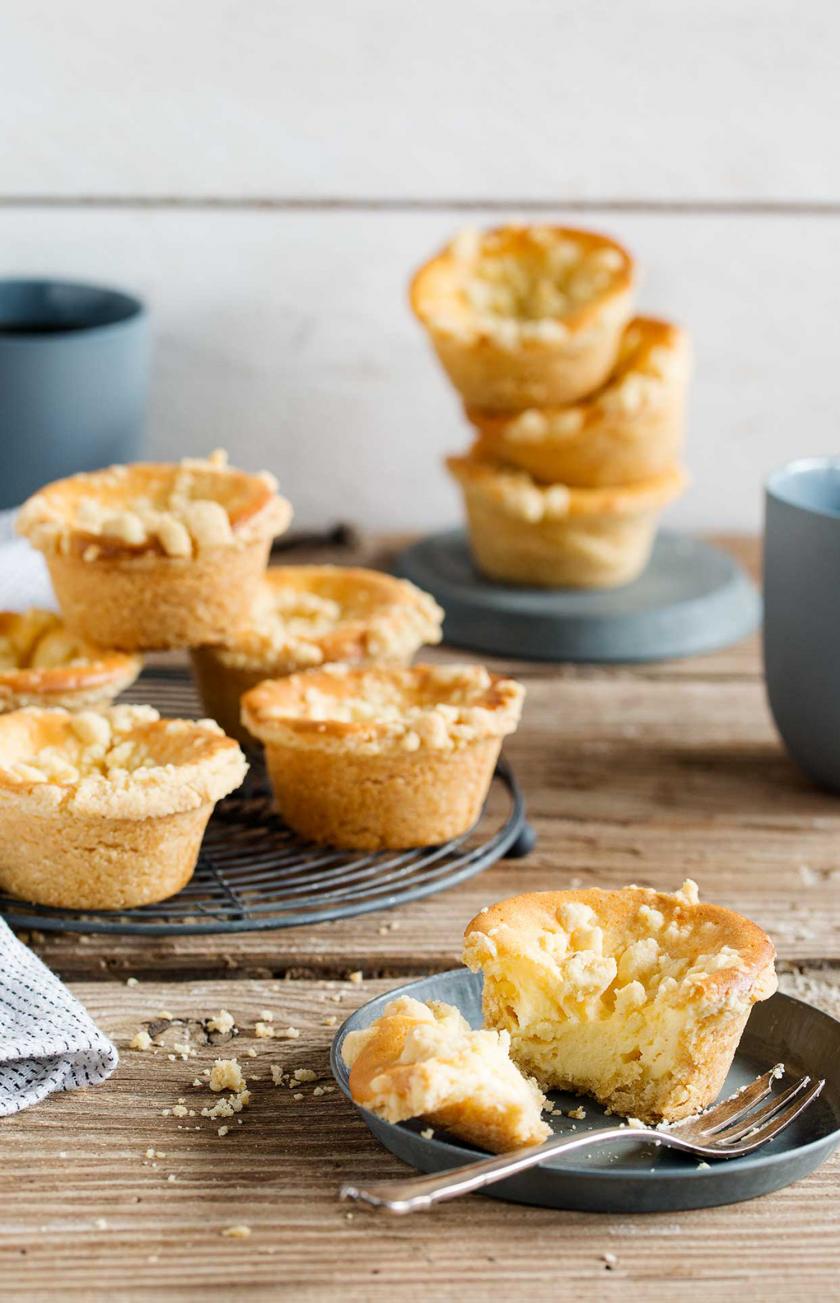 Mehrere Käsekuchemuffins mit Streuseln stehen auf einem Kuchengitter und auf einem Teller.