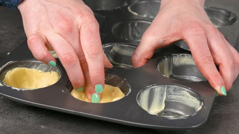 Der Mürbeteig für den Boden der Käsekuchenmuffins mit Streuseln wird in die Muffinförmchen gedrückt.