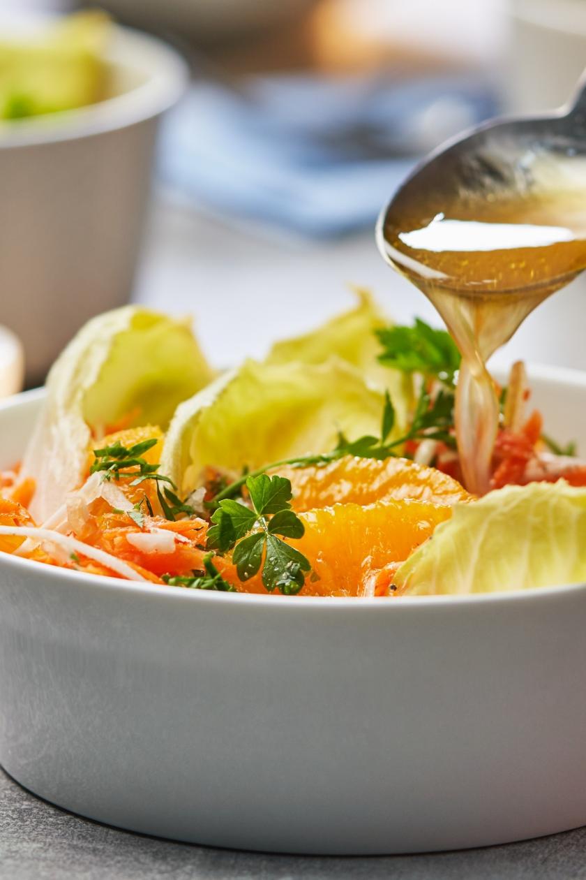 Karottensalat mit Sellerie, Äpfeln und Orangenfilets in einer Schale, wird mit Dressing übergossen.