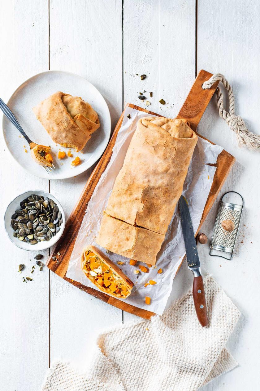 Kürbisstrudel angeschnitten auf einem Holzbrett, daneben ein Teller mit einem Stück Kürbisstrudel und eine Schale Kürbiskerne.