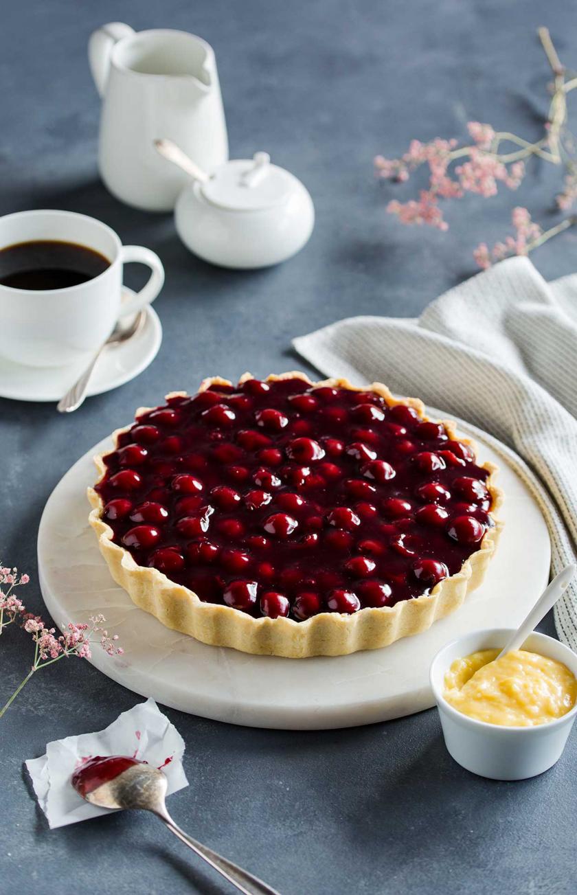 Lemon Curd Tarte mit Balsamico-Kirschen liegt auf einem Kuchenteller und steht auf einem gedeckten Tisch.