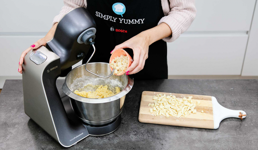 Weiße Schokolade wird zum teig der Macadamia Cookies in die Rührschüssel der Küchenmaschine gegeben.