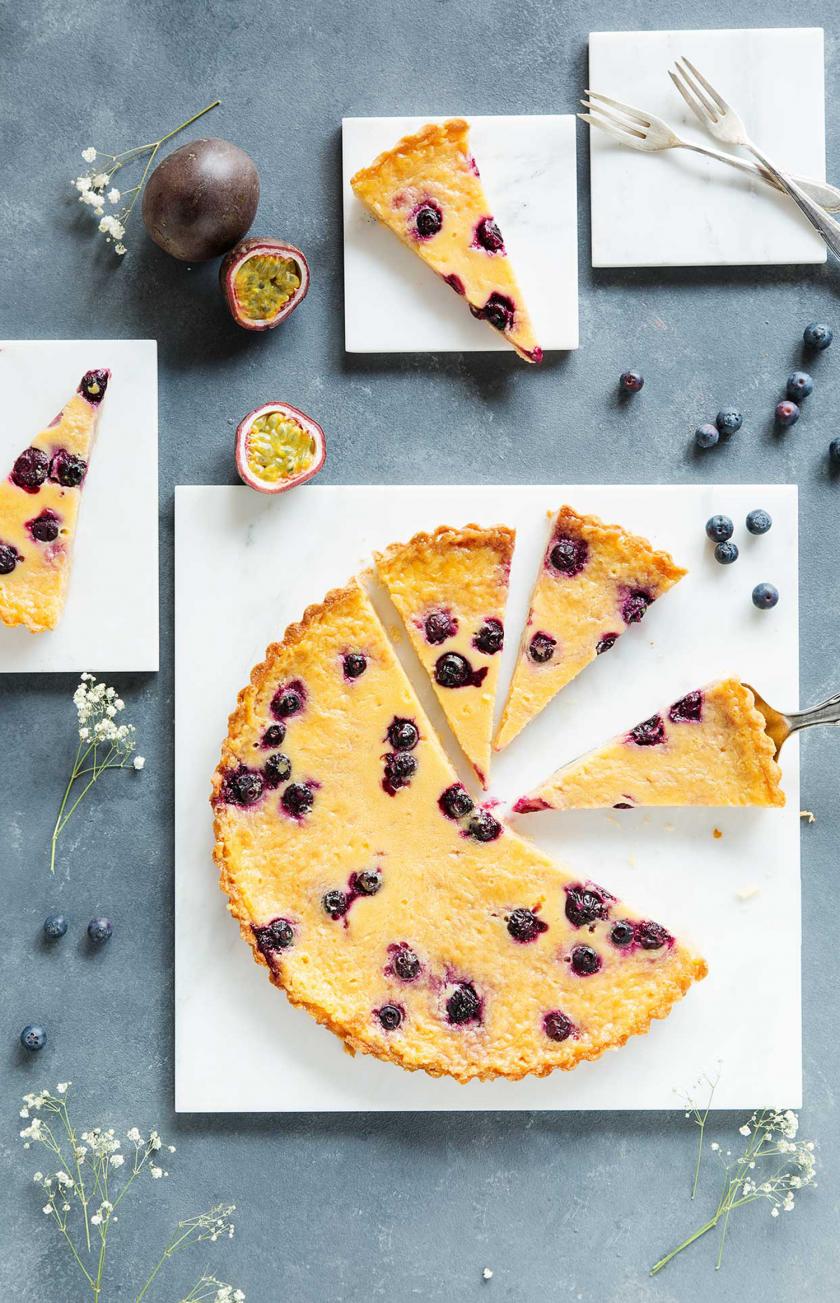 Maracuja-Tarte mit Blaubeeren angeschnitten auf einer Kuchenplatte.