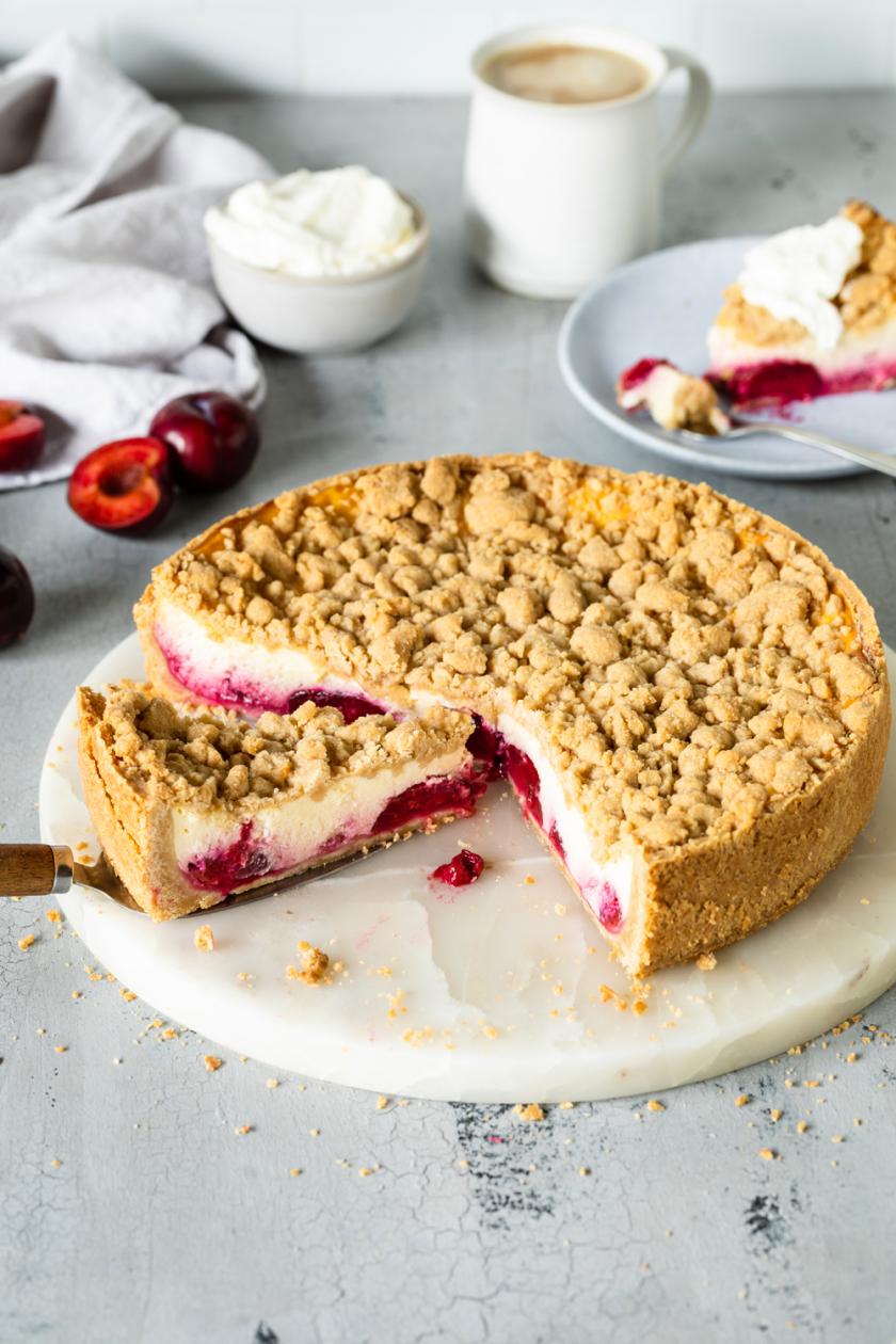 Pflaumenkuchen mit Pudding und Zimtstreuseln angeschnitten auf einer Kuchenplatte, daneben frische Pflaumen