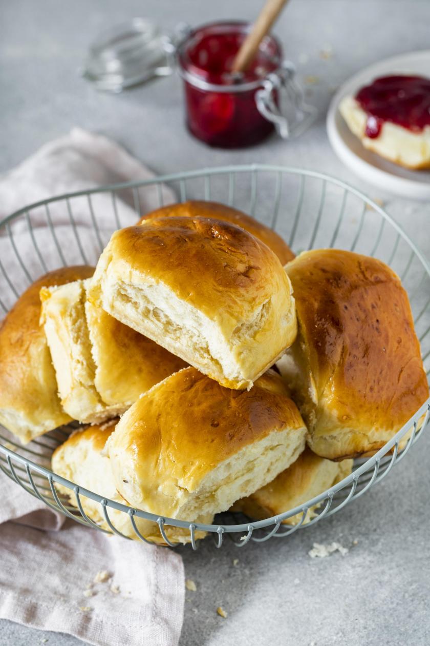 Milchbrötchen in einem Brötchenkorb aus Draht, daneben rote Marmelade auf einem Tisch