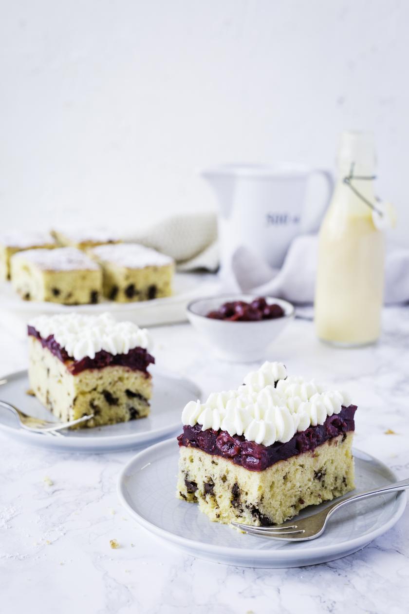 Zwei Stücke Eierlikörkuchen mit Sahne und Kirschen auf Tellern.