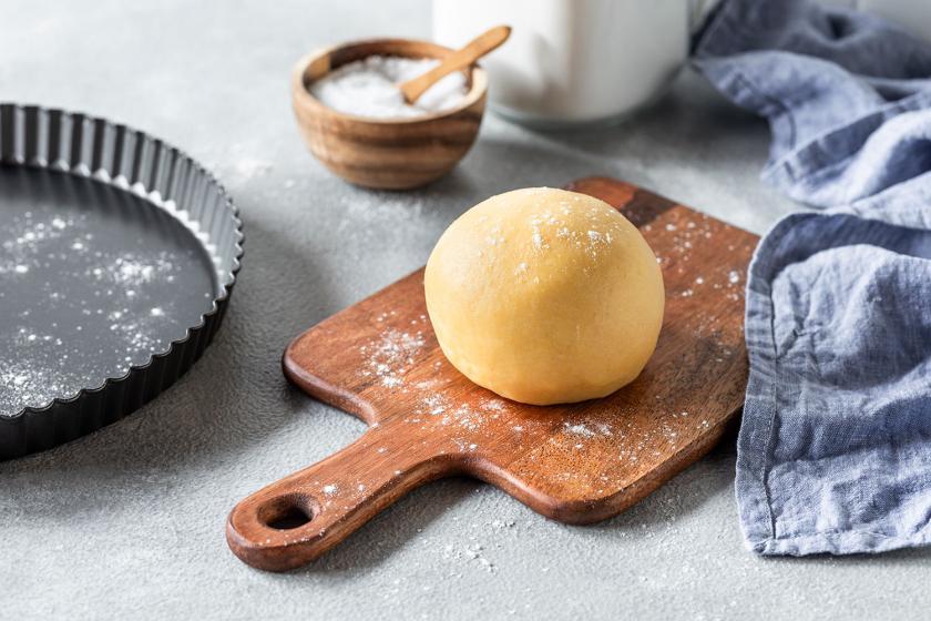 Quicheteig liegt als Kugel geformt auf einem Holzbrett, daneben Zutaten wie Mehl