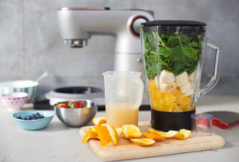 Für die Smoothie Bowl mit Bananen liegen Orangen geschnitten auf einem Holzbrett. Daneben ein Mixer mit allen Zutaten gefüllt.