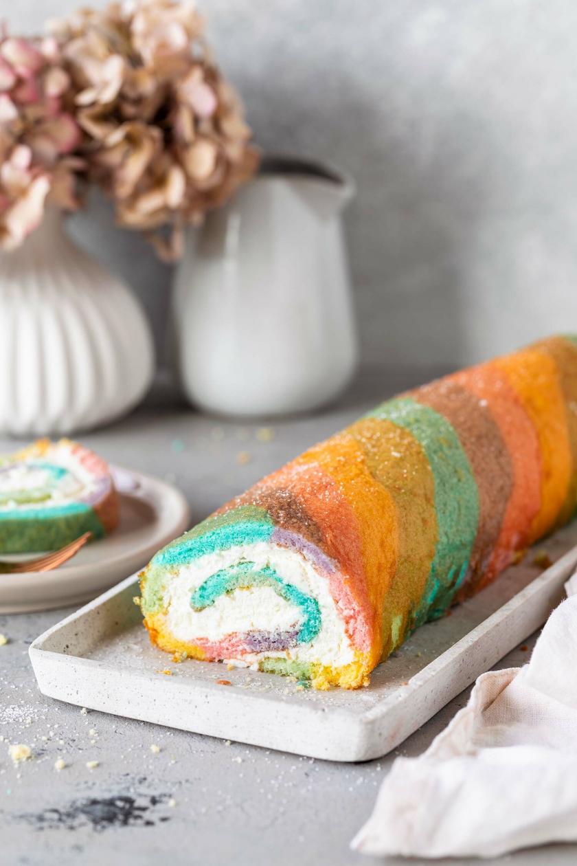 Regenbogen Biskuitrolle auf einer Tortenplatte, dahinter Blumen auf einem Tisch