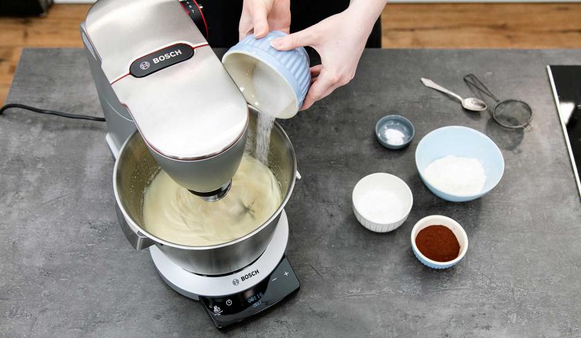 Für den Boden der Schoko-Bananen-Torte wird Zucker zur Eimasse in die Rührschüssel der Küchenmaschine gegeben.