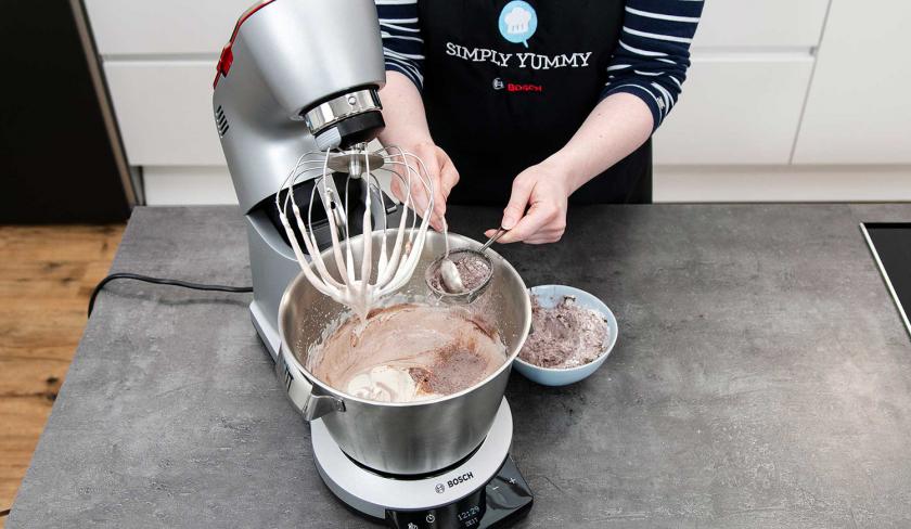 Mehl wird auf die Eimasse in der Rührschüssel der Küchenmaschine gesiebt.