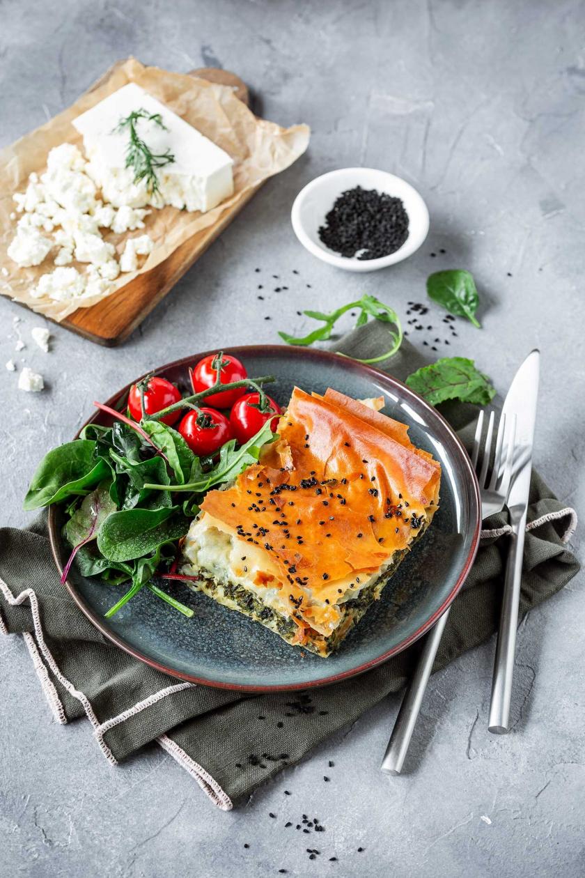Ein Stück Spanakopita auf einem Teller mit Salat und Tomaten.