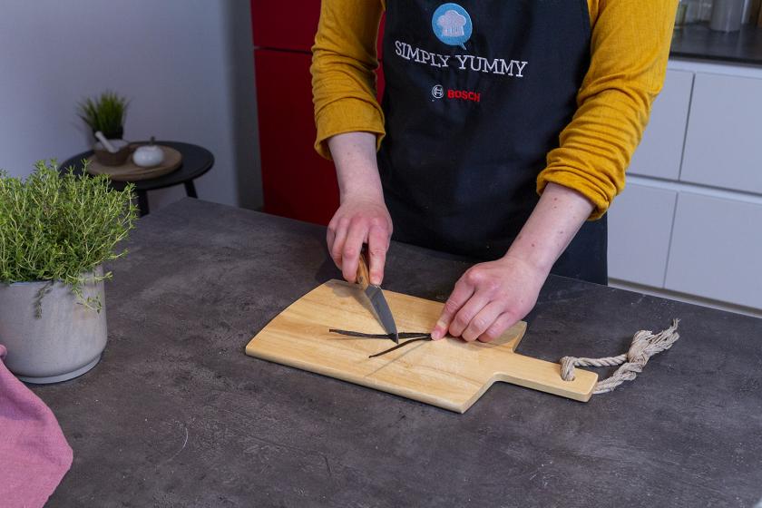 Eine Vanilleschote wird mit einem Messer auf einem Holzbrett ausgekratzt.