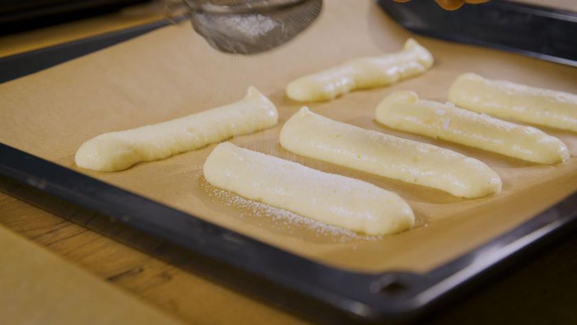 Die rohen Löffelbiskuits liegen auf einem Backblech mit Backpapier.