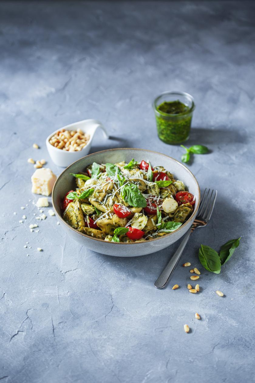 Tortellinisalat mit Pesto, Mozzarella, Tomaten und Basilikum auf einem Teller mit angelegter Gabel sowie Pinienkernen und Pesto.