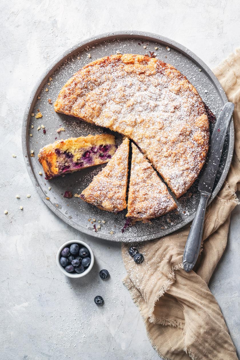 Zitronen-Blaubeer-Kuchen mit Ricotta angeschnitten auf einer Kuchenplatte.