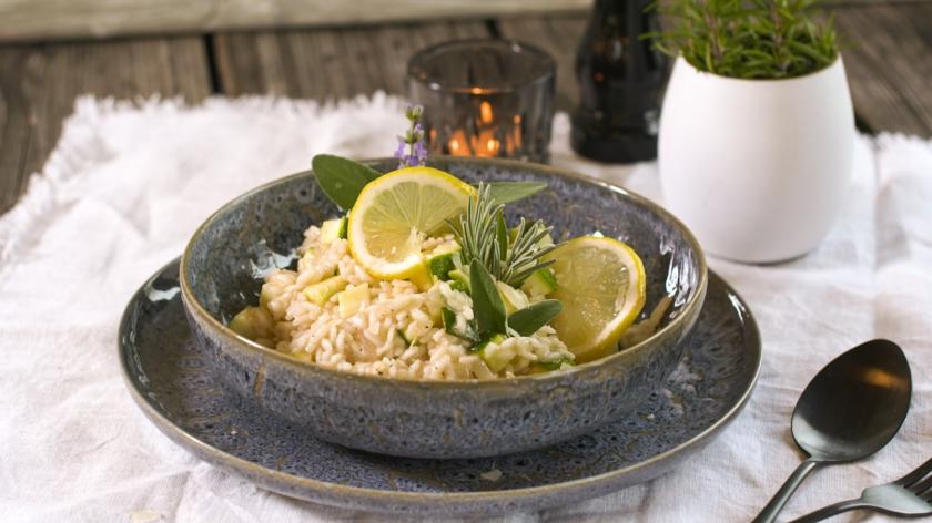 Zucchini-Risotto mit Zitronen dekoriert in dunkler Schale auf gedecktem Tisch.