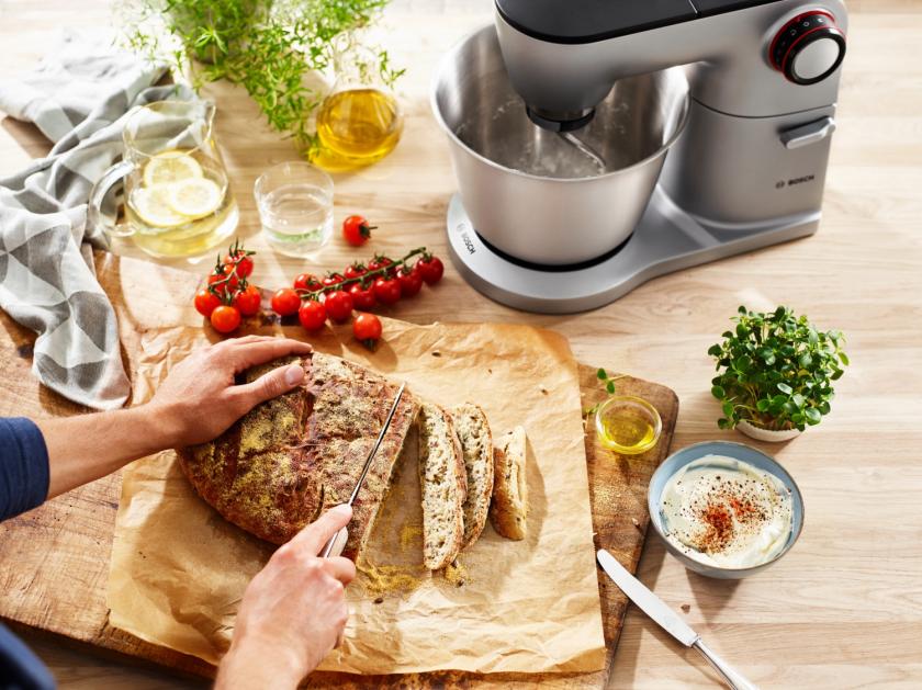 Küchenmaschine OptiMUM im Hintergrund, Frau schneidet Brot im Vordergrund.